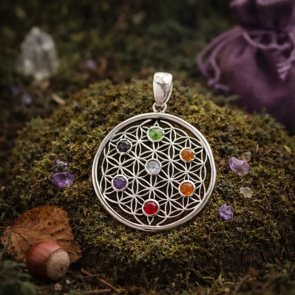 Pendentif rond en argent représentant une Fleur de Vie finement ciselée et sertie de 7 petites pierres naturelles colorées, posé sur une souche moussue en forêt.