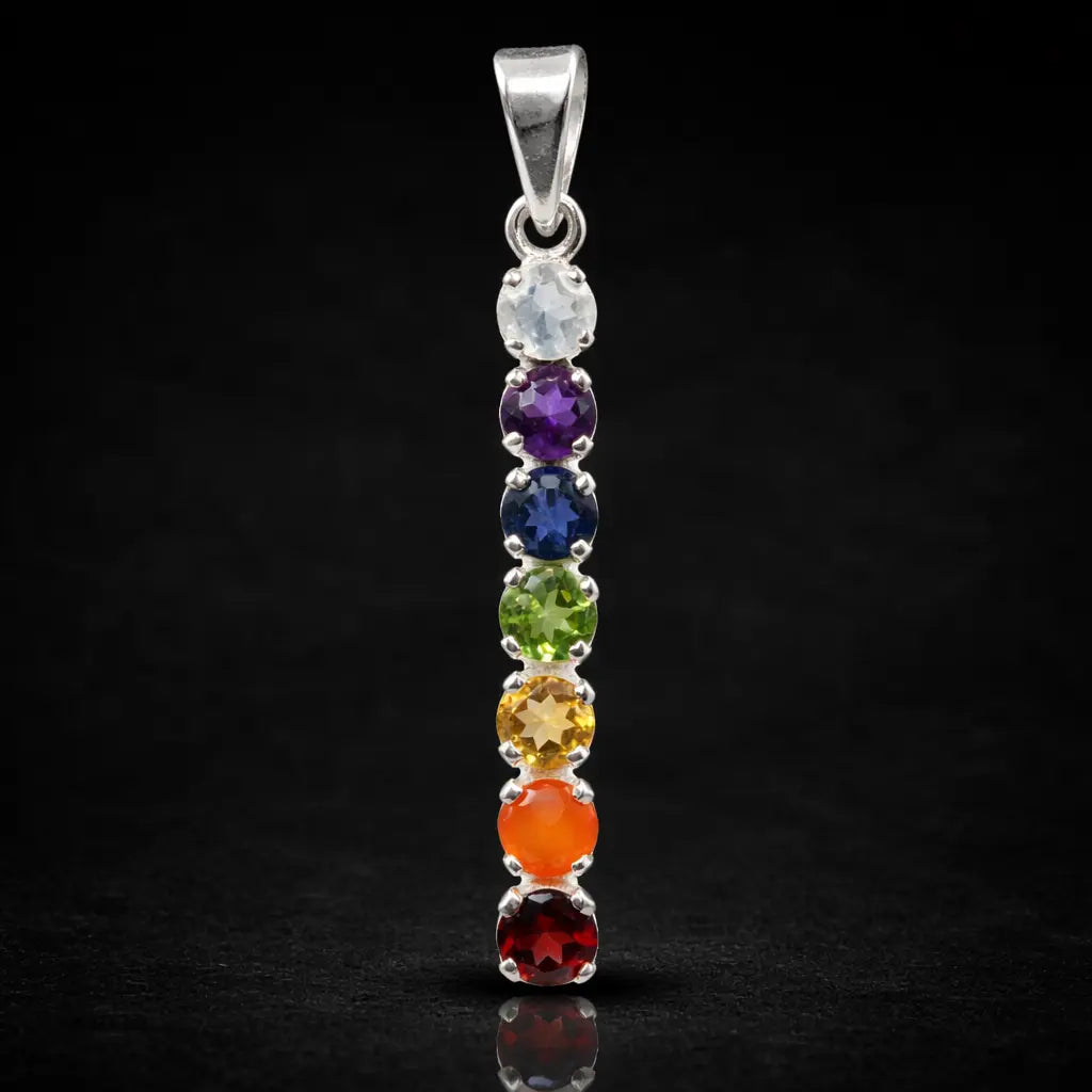 Un pendentif en argent composé de sept pierres naturelles taillées et alignées verticalement aux couleurs des chakras, présenté avec son reflet sur un fond noir.