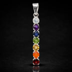 Un pendentif en argent composé de sept pierres naturelles taillées et alignées verticalement aux couleurs des chakras, présenté avec son reflet sur un fond noir.