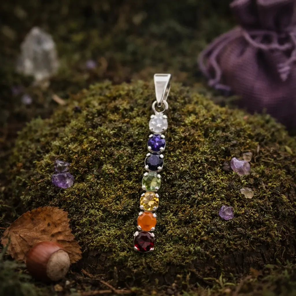 Pendentif vertical en argent serti de 7 pierres naturelles rondes aux couleurs des chakras, posé sur un lit de mousse forestière avec un pochon violet.