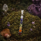 Pendentif vertical en argent serti de 7 pierres naturelles rondes aux couleurs des chakras, posé sur un lit de mousse forestière avec un pochon violet.