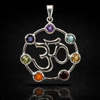 Un pendentif Om en argent au centre d'un cercle serti de sept pierres naturelles taillées aux couleurs des chakras, présenté avec son reflet sur un fond noir.