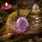 Pendentif ovale en améthyste polie aux nuances violettes translucides, monté sur un cordon noir, posé sur une tranche de bois moussue avec une bougie violette et des cristaux.