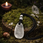 Pendentif de vertu en cristal de roche translucide de forme goutte, orné de deux perles cristallines, posé sur une souche moussue avec une bougie allumée en arrière-plan.