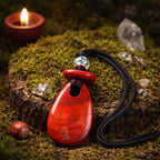 Pendentif de vertu en jaspe rouge poli de forme goutte allongée, surmonté d'une perle translucide et d'une petite pièce de jaspe, posé sur une souche moussue.