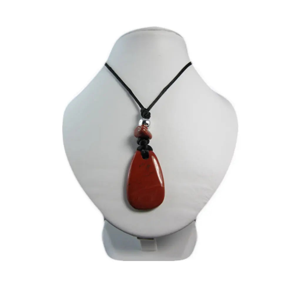 Un pendentif en jaspe rouge taillé en forme de goutte polie, agrémenté d'une petite perle argentée et d'une pierre roulée, présenté sur un buste d'exposition blanc.