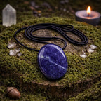 Pendentif en sodalite ovale polie bleu nuit avec des veines blanchâtres, monté sur un cordon noir et posé sur une souche moussue avec une bougie et un cristal de roche.