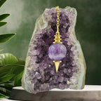 Pendule Séphoroton en améthyste violette avec monture et chaîne en métal doré, suspendu devant une géode d'améthyste dans un décor zen avec des feuilles vertes.