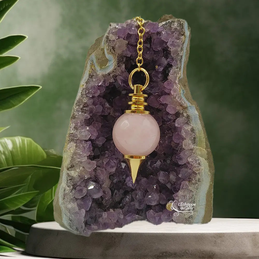 Pendule Séphoroton en quartz rose avec monture et chaîne en métal doré, suspendu devant une géode d'améthyste violette dans un décor zen agrémenté de feuilles vertes.