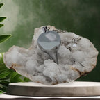 Pendule goutte en métal argenté brillant, posé sur une géode de cristaux blancs dans un décor zen.