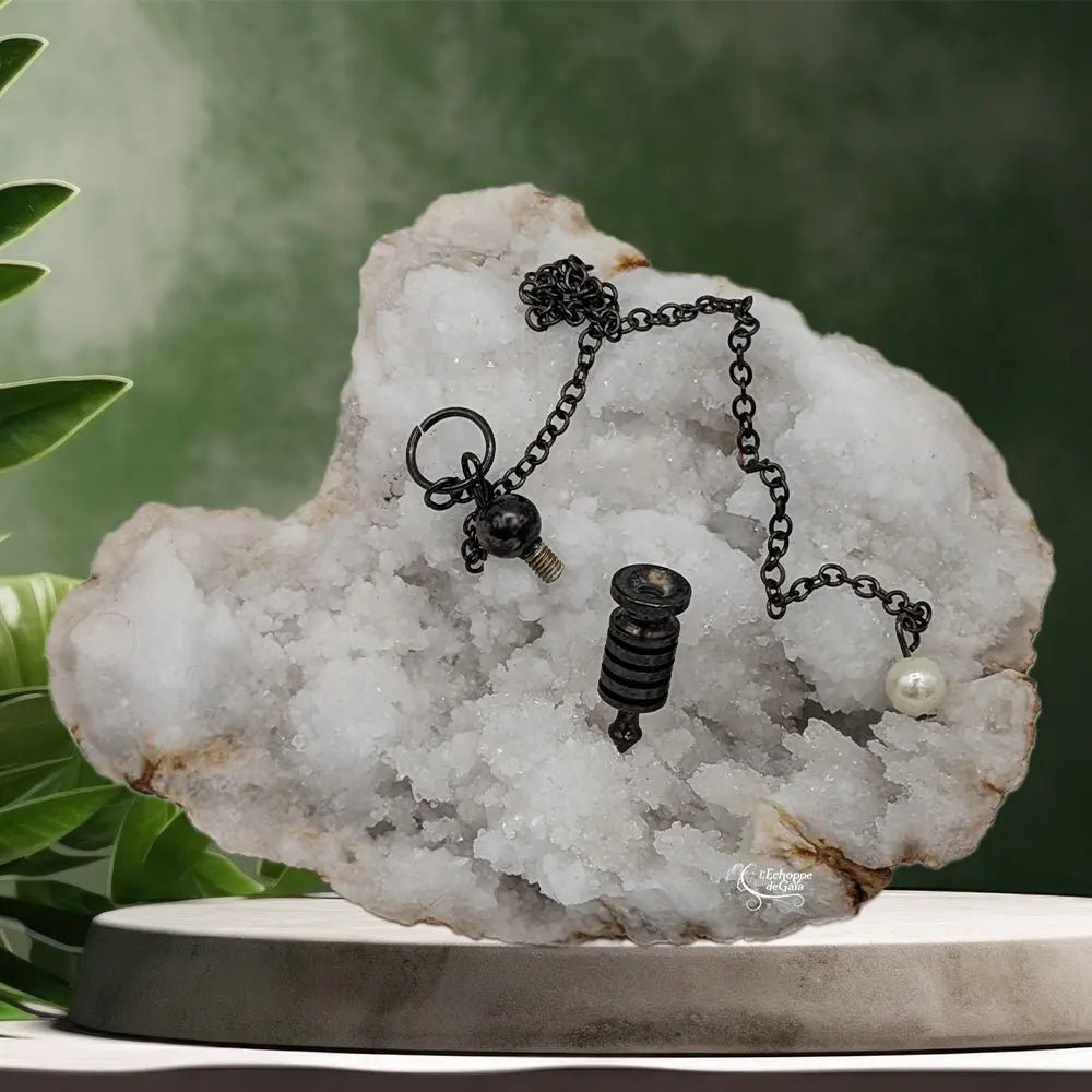 Pendule Isis noir à témoin dévissé révélant sa cavité interne posé sur un cristal de roche blanc.