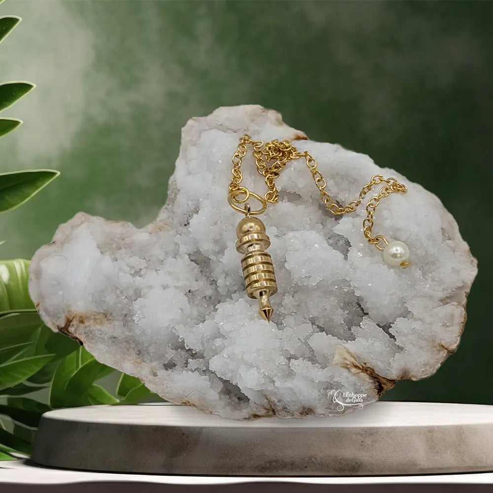 Pendule Isis doré en métal brillant suspendu devant un cristal de roche blanc brut et décor vert.