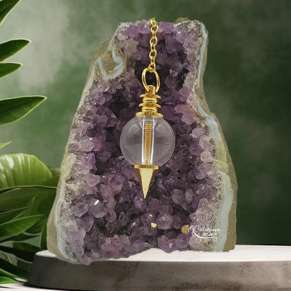 Pendule Séphoroton en cristal de roche et métal doré avec spirale interne, suspendu devant une géode d'améthyste.