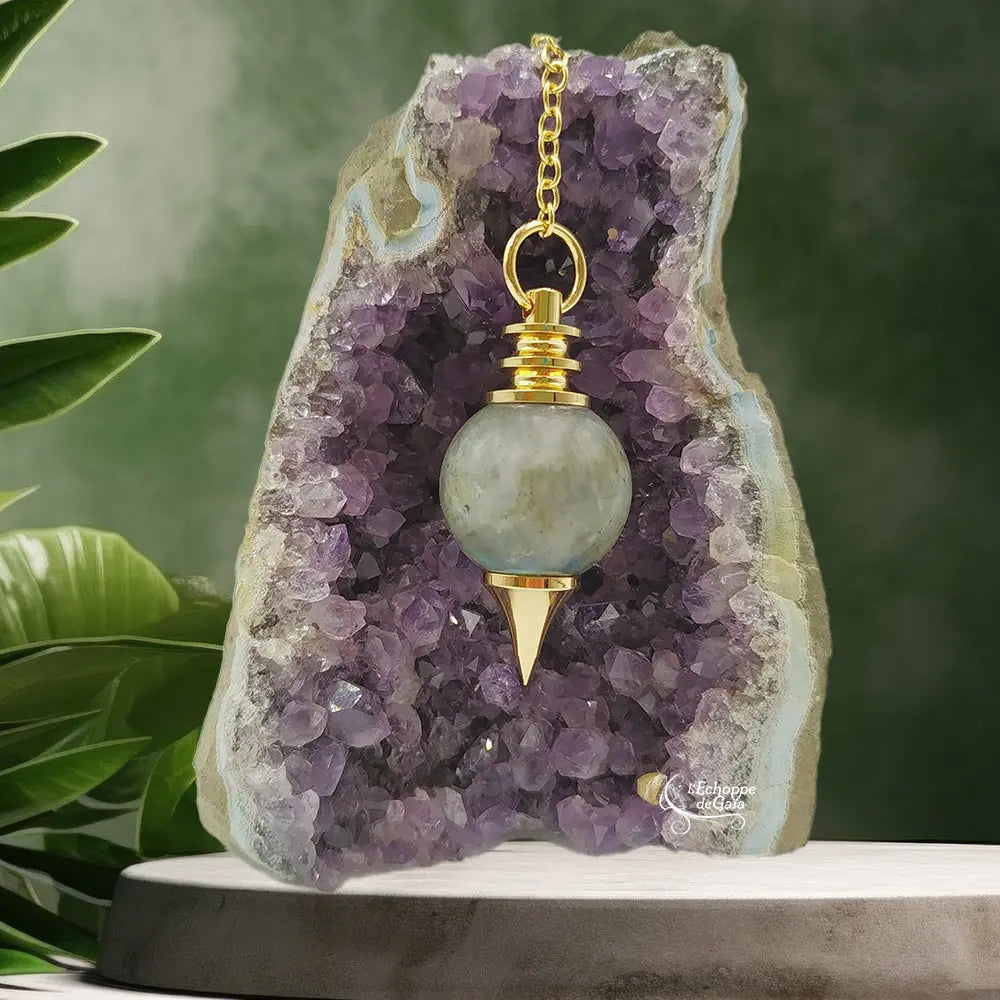 Pendule Séphoroton en labradorite aux reflets bleutés avec monture et chaîne en métal doré, suspendu devant une géode d'améthyste violette dans un décor zen agrémenté de feuilles vertes.