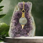 Pendule Séphoroton en labradorite aux reflets bleutés avec monture et chaîne en métal doré, suspendu devant une géode d'améthyste violette dans un décor zen agrémenté de feuilles vertes.