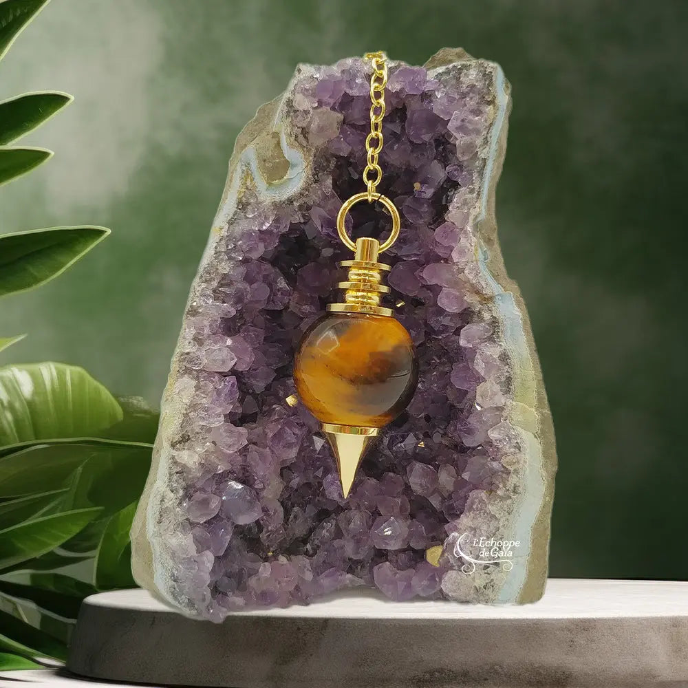 Pendule Séphoroton en œil de tigre avec monture et chaîne en métal doré, suspendu devant une géode d'améthyste violette dans un décor zen agrémenté de feuilles vertes.