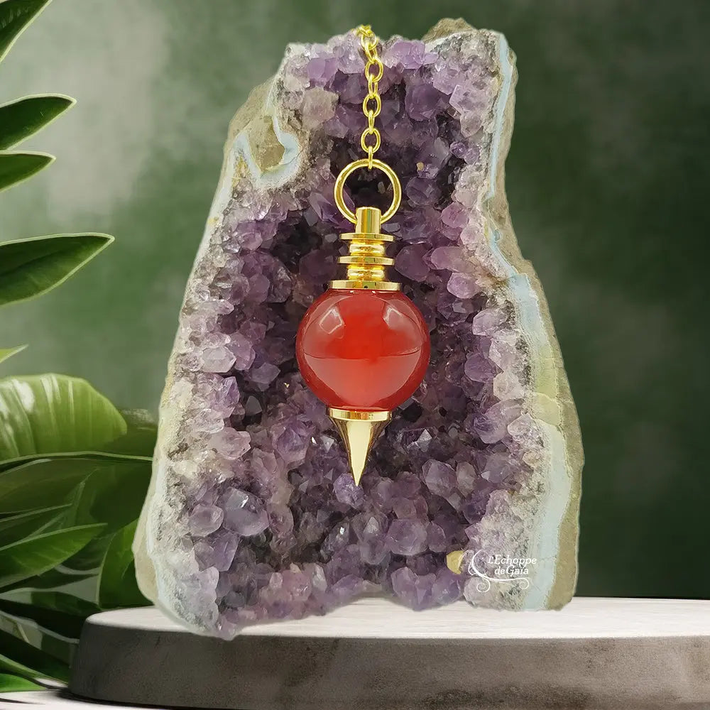 Pendule Séphoroton en cornaline rouge-orangé avec monture et chaîne en métal doré, suspendu devant une géode d'améthyste violette dans un décor zen agrémenté de feuilles vertes.