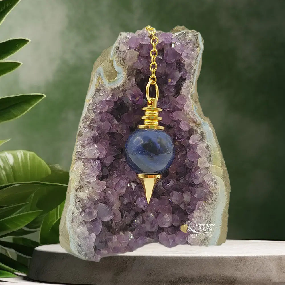 Pendule Séphoroton en sodalite bleu foncé et métal doré, suspendu par sa chaîne devant une géode d'améthyste violette dans un décor zen avec des feuilles vertes.