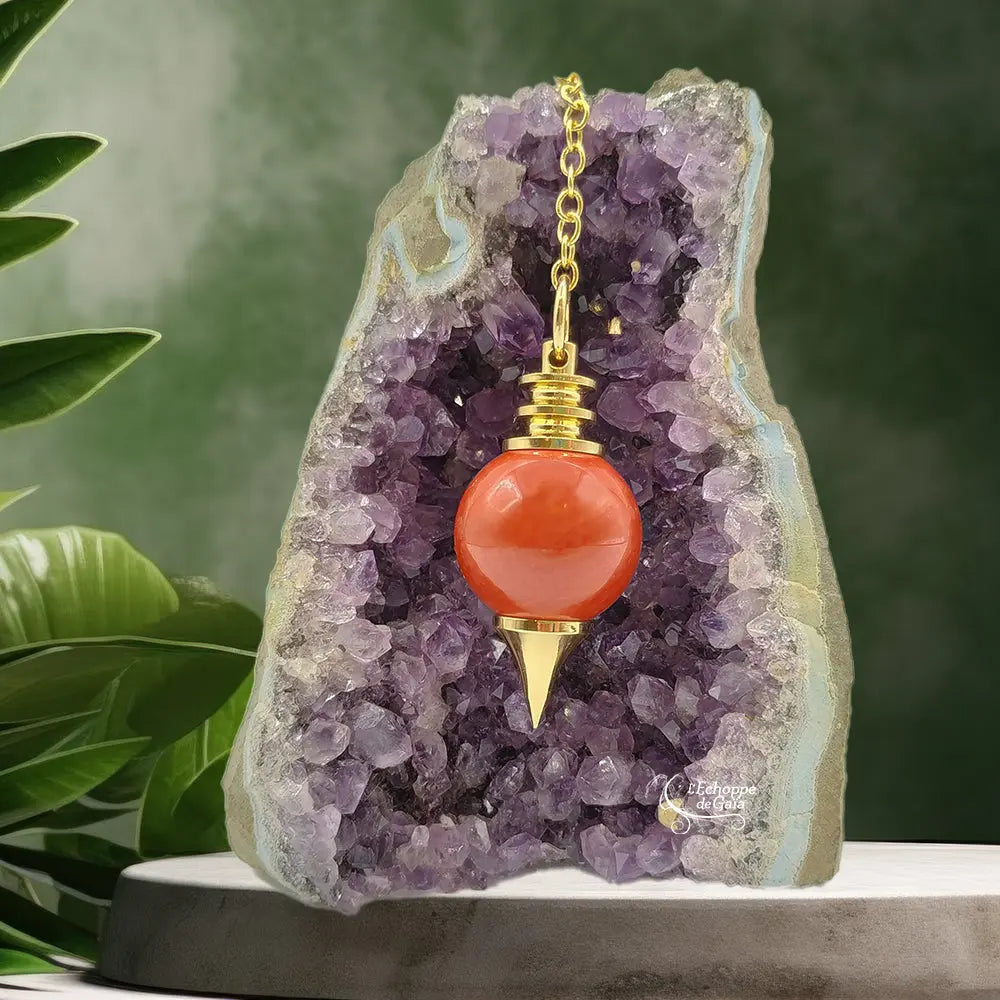 Pendule Séphoroton en jaspe avec monture et chaîne en métal doré, suspendu devant une géode d'améthyste violette dans un décor zen avec des feuilles vertes.