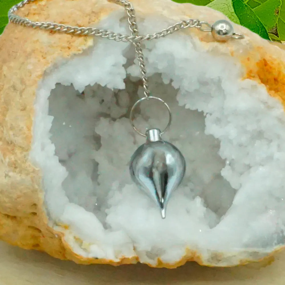 Pendule en métal chromé brillant en forme de goutte, suspendu par sa chaîne devant l'ouverture d'une géode de cristaux blancs.