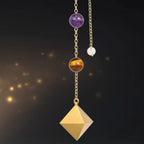 Pendule Lumière Fangain en métal doré brossé de forme pyramidale, suspendu par sa chaîne ornée de perles d'améthyste et d'œil de tigre sur un fond sombre scintillant.
