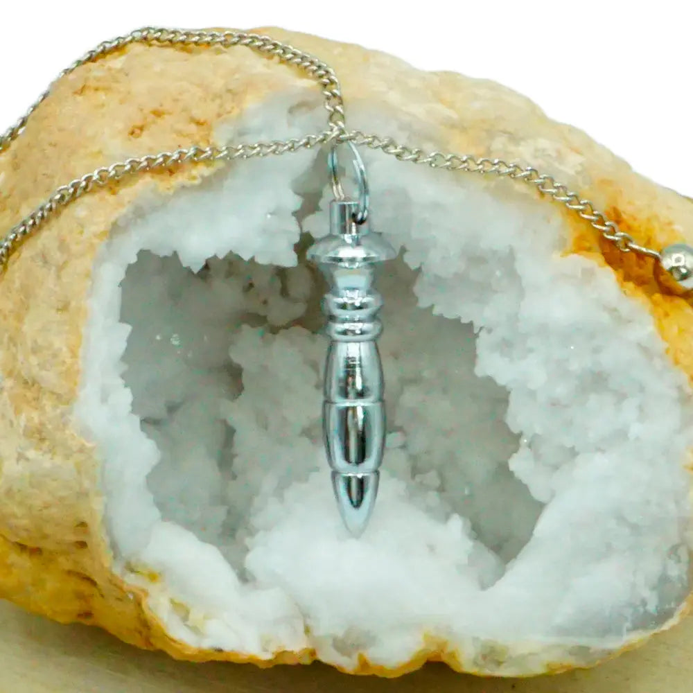 Pendule égyptien du Nil en métal argenté brillant, suspendu par sa chaîne devant l'ouverture d'une géode de cristaux blancs.