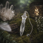 Pendule égyptien Thot en cristal de roche transparent sculpté, posé sur une écorce moussue à côté d'un amas de quartz, d'une plume blanche et d'un photophore.