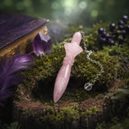 Pendule égyptien en quartz rose posé sur une souche de bois moussue, à côté d'un grimoire violet, d'une pointe d'améthyste et d'une plume violette.