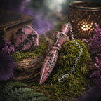 Pendule égyptien Thot en rhodonite rose marbrée de noir, posé sur un lit de mousse sylvestre à côté d'un bloc de pierre brute, d'un vieux grimoire et d'un photophore métallique ajouré.