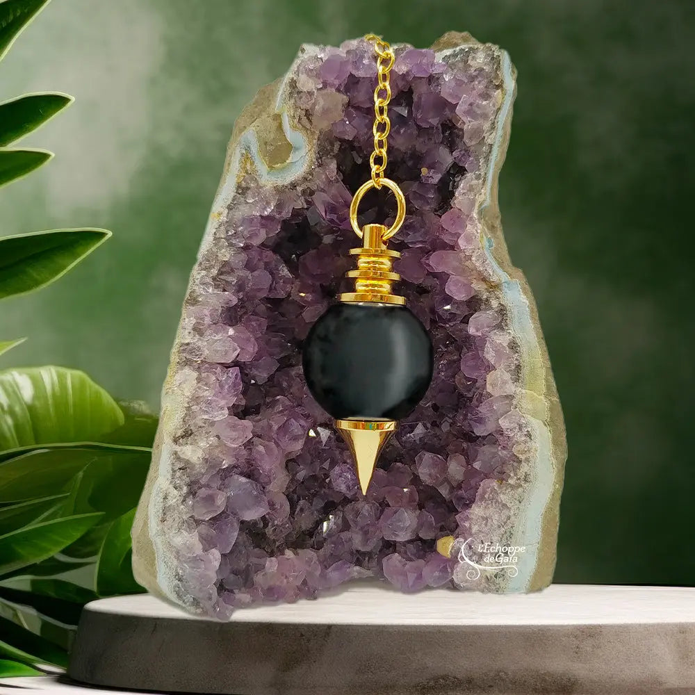 Pendule Séphoroton en obsidienne noire et métal doré, suspendu par sa chaîne devant une géode d'améthyste violette dans un décor zen avec des feuilles vertes.
