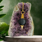 Pendule Séphoroton en obsidienne noire et métal doré, suspendu par sa chaîne devant une géode d'améthyste violette dans un décor zen avec des feuilles vertes.