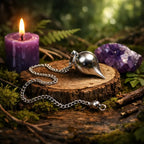 Pendule en forme de goutte en métal chromé brillant posé sur un socle de bois avec une améthyste et une bougie violette en arrière-plan.