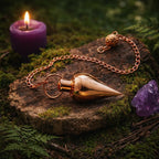 Pendule en métal cuivré en forme de goutte déposé sur une souche de bois moussue à côté d'une bougie et d'un cristal.