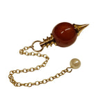 Pendule Séphoroton en cornaline orange avec monture et chaîne en métal doré, terminé par une perle nacrée sur fond blanc.