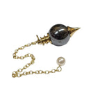 Pendule Séphoroton en hématite gris métallique et métal doré avec chaîne et perle terminale nacrée sur fond blanc.