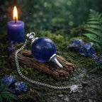Pendule Sephoroton en Lapis Lazuli bleu profond avec éclats de pyrite et monture argentée, posé sur un bois ancien avec une bougie violette et des pierres brutes.