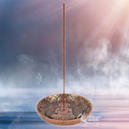 Porte-encens rond en métal effet bronze avec motif Bouddha au centre, bâton d’encens planté verticalement, sur fond brumeux.