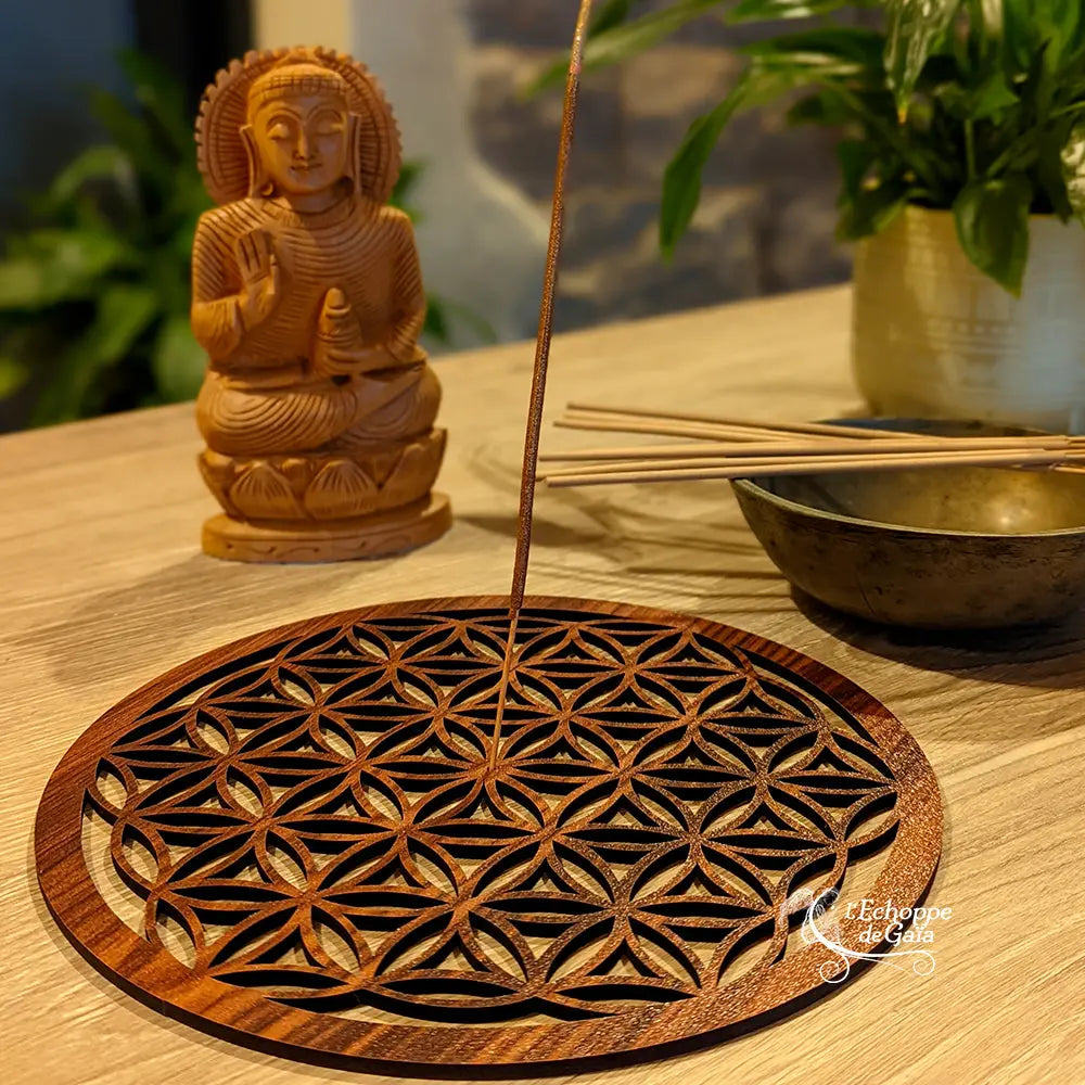 Porte-encens rond en bois ajouré motif Fleur de Vie, avec bâton d’encens planté, sur une table près d’une statue de Bouddha et de bâtonnets d’encens.