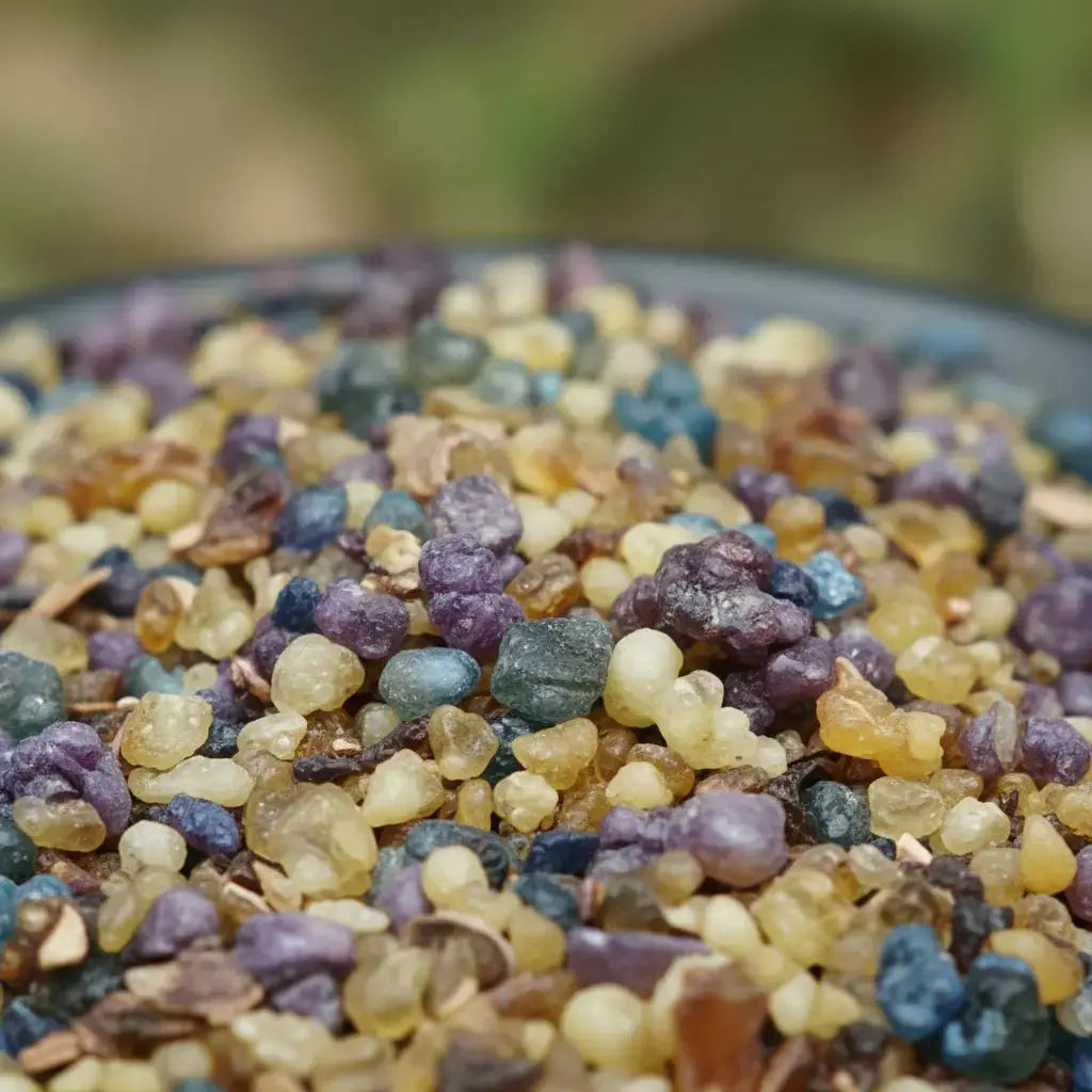 Gros plan sur des grains d’encens Saint Antoine multicolores dans des tons violets, bleus et dorés sur fond flou.
