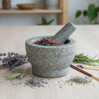 Mortier en pierre rempli de grains d’encens Saint Jean Baptiste multicolores, entouré de lavande et d’herbes sur table en bois claire.
