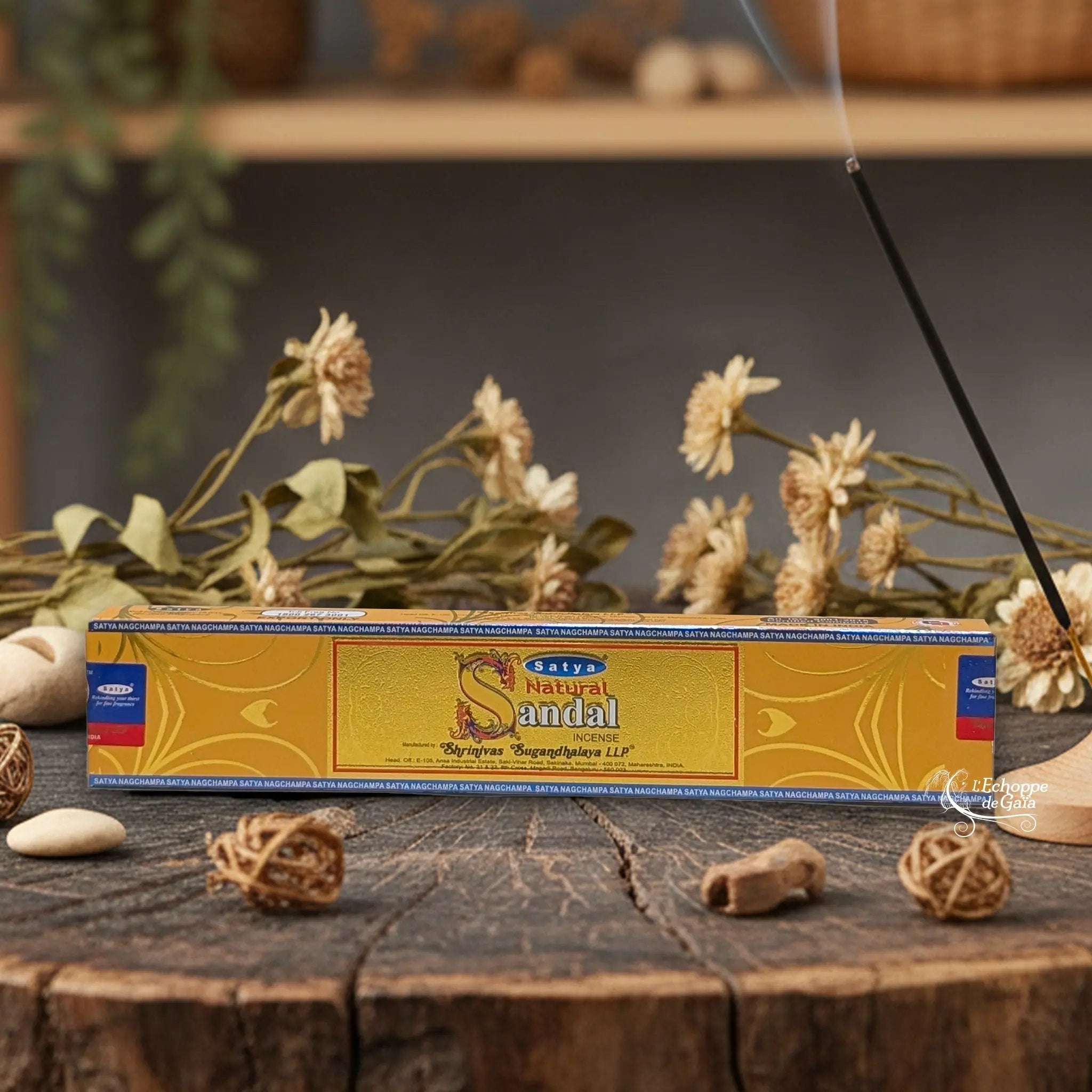 Boîte d'encens Satya "Natural Sandal" posée sur une table en bois avec des fleurs séchées et un bâton d'encens fumant.