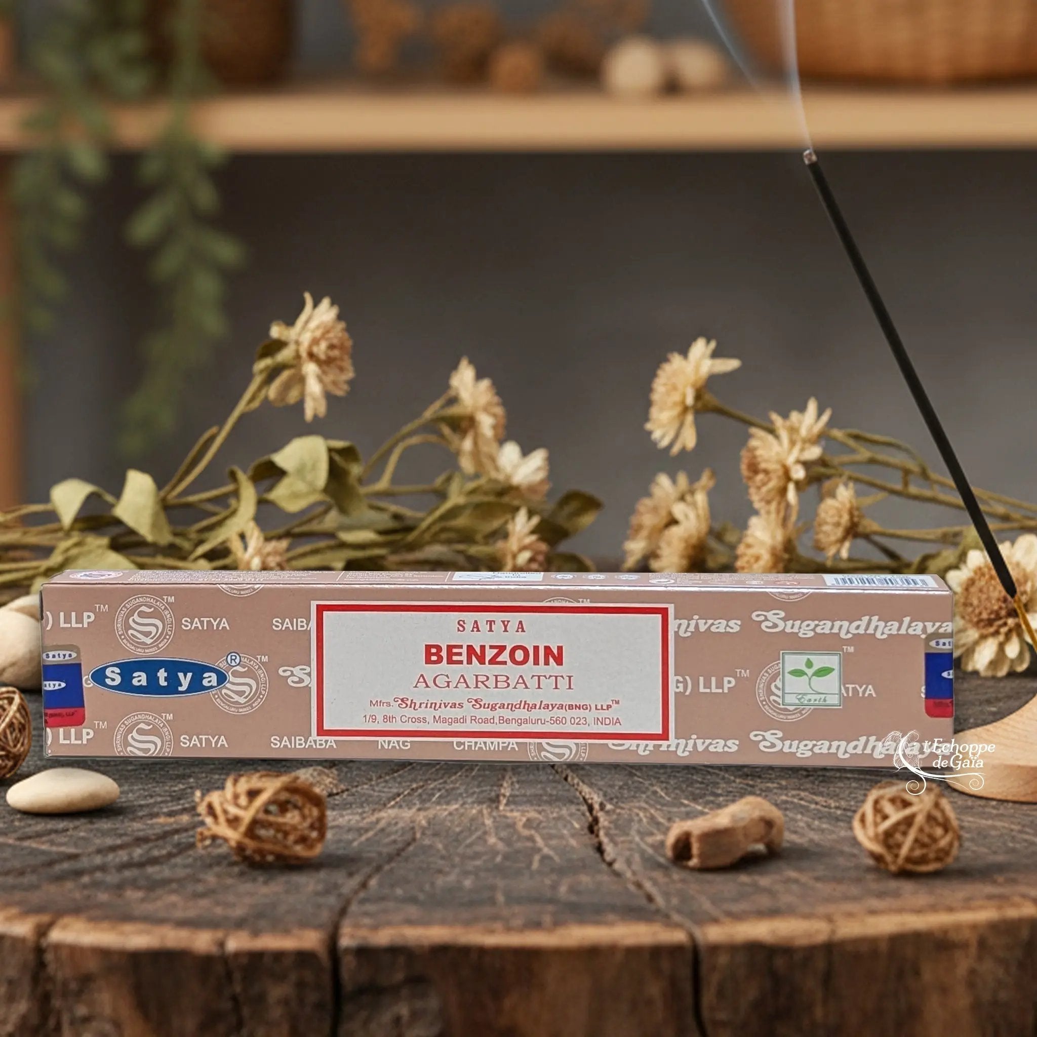 Boîte d'encens Satya "Benzoin" Agarbatti posée sur une table en bois avec des fleurs séchées et un bâton d'encens fumant.