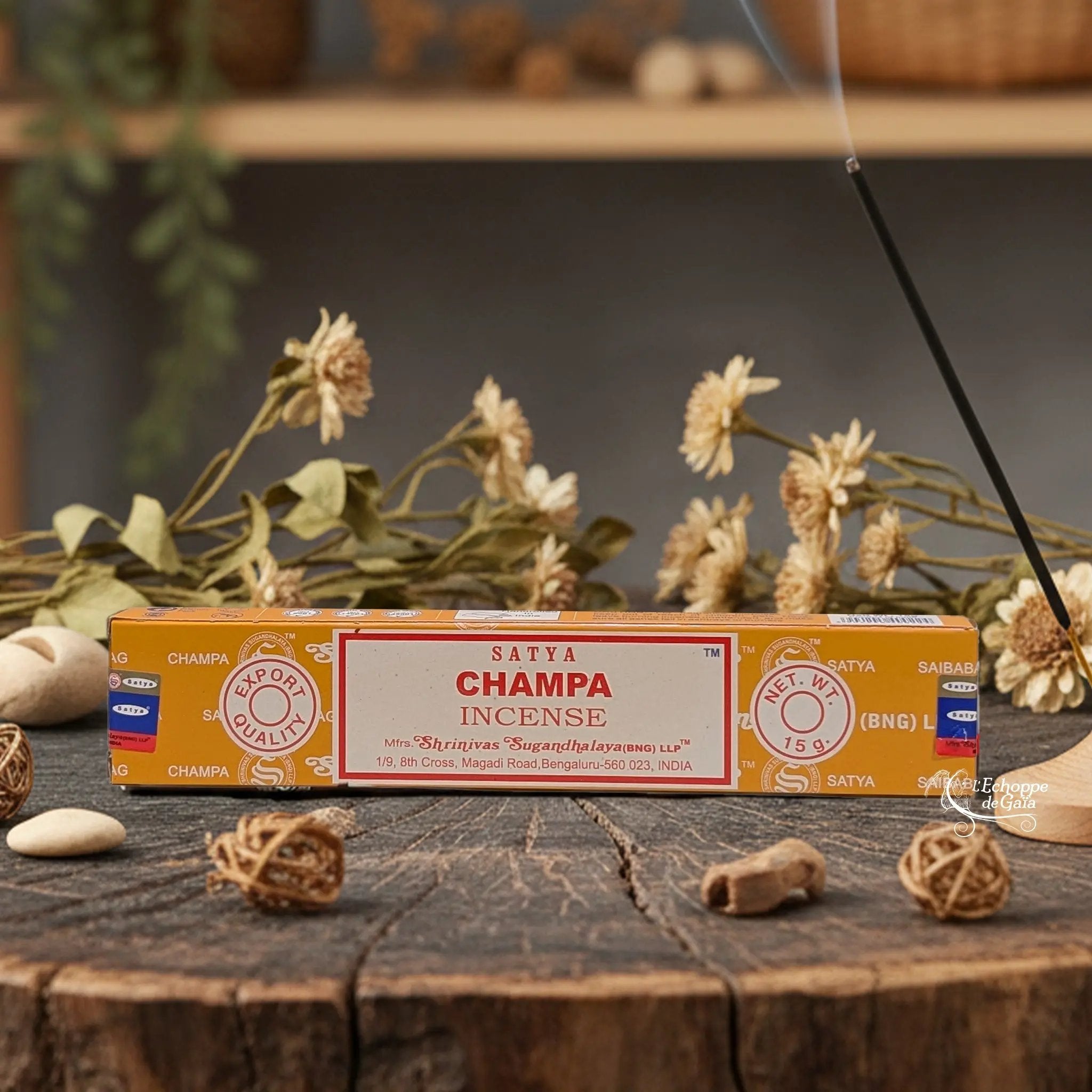 Boîte d'encens Satya "Champa" posée sur une table en bois avec des fleurs séchées et un bâton d'encens fumant.