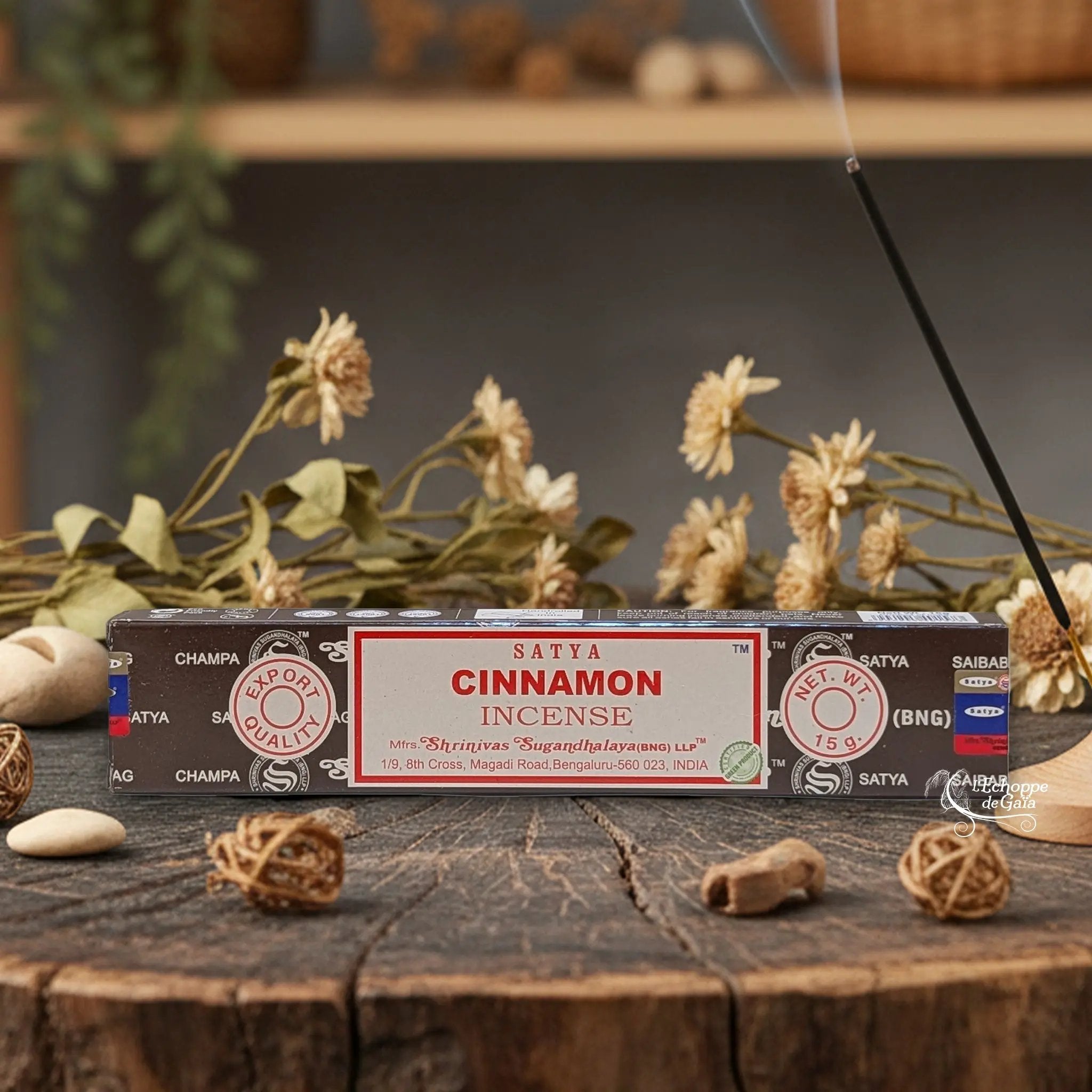 Boîte d'encens Satya "Cinnamon" posée sur une table en bois avec des fleurs séchées et un bâton d'encens fumant.