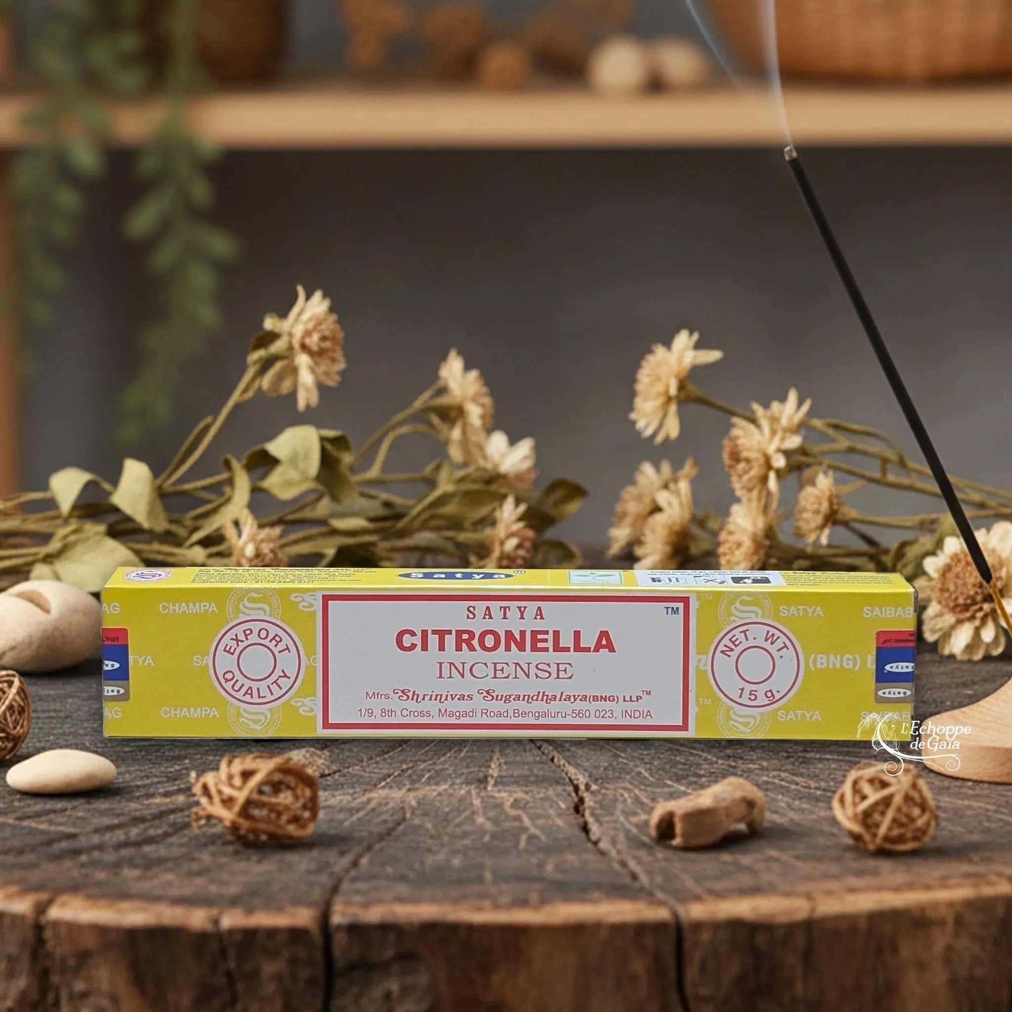 Boîte d'encens Satya "Citronella" posée sur une table en bois avec des fleurs séchées et un bâton d'encens fumant.