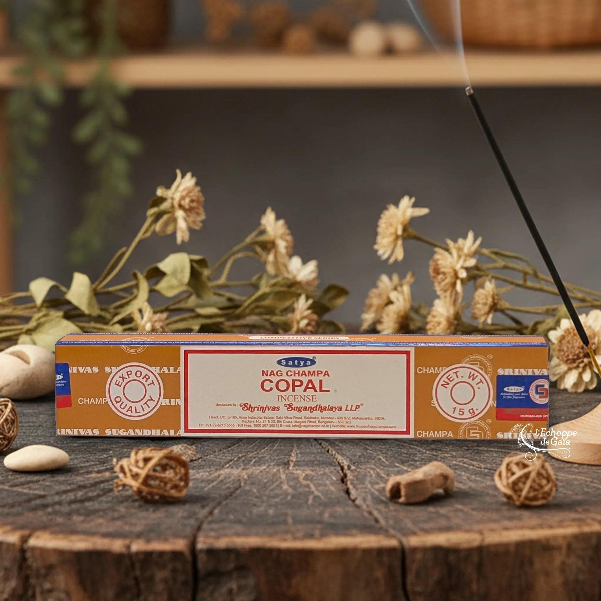 Boîte d'encens Satya Nag Champa "Copal" posée sur une table en bois avec des fleurs séchées et un bâton d'encens fumant.