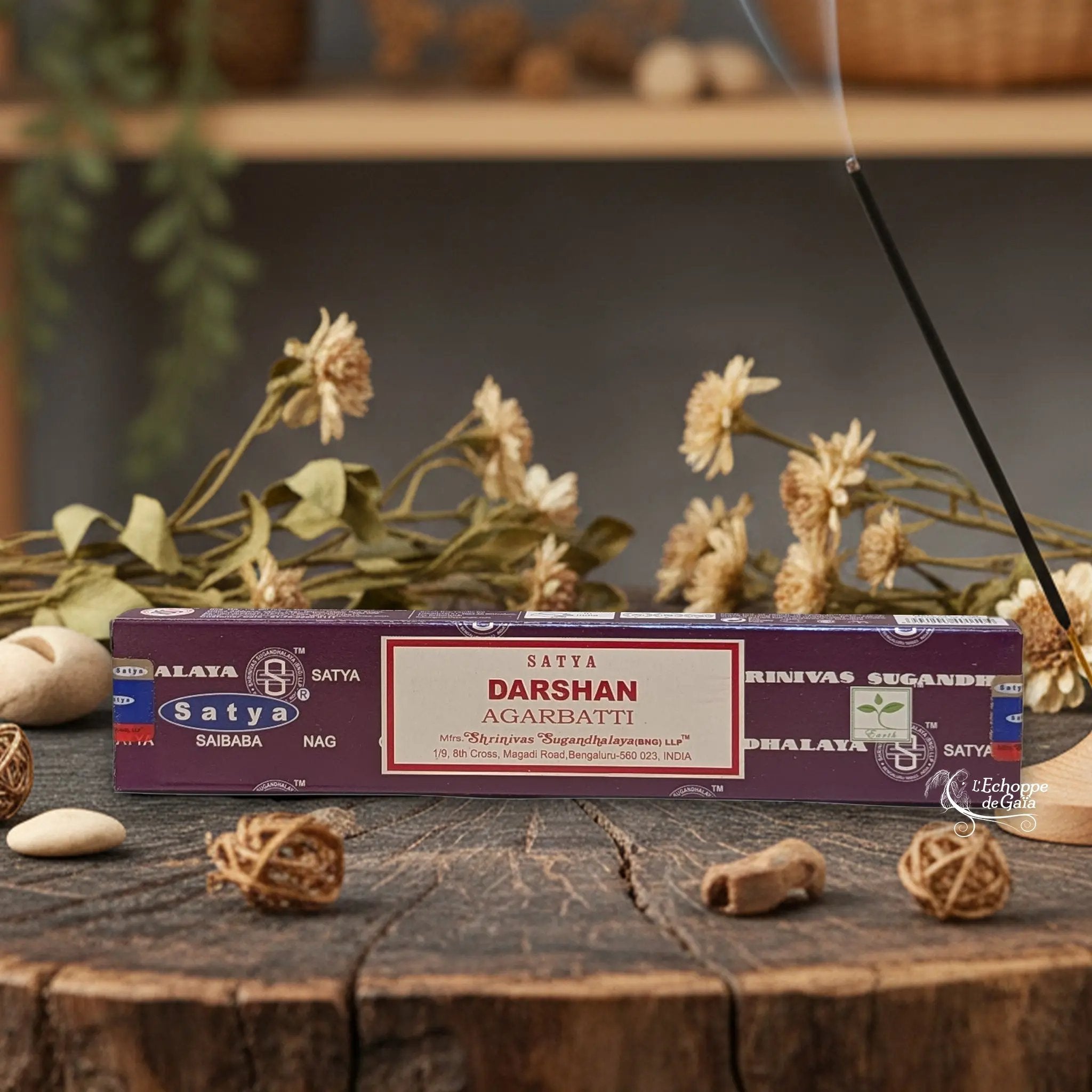 Boîte d'encens Satya "Darshan" Agarbatti posée sur une table en bois avec des fleurs séchées et un bâton d'encens fumant.