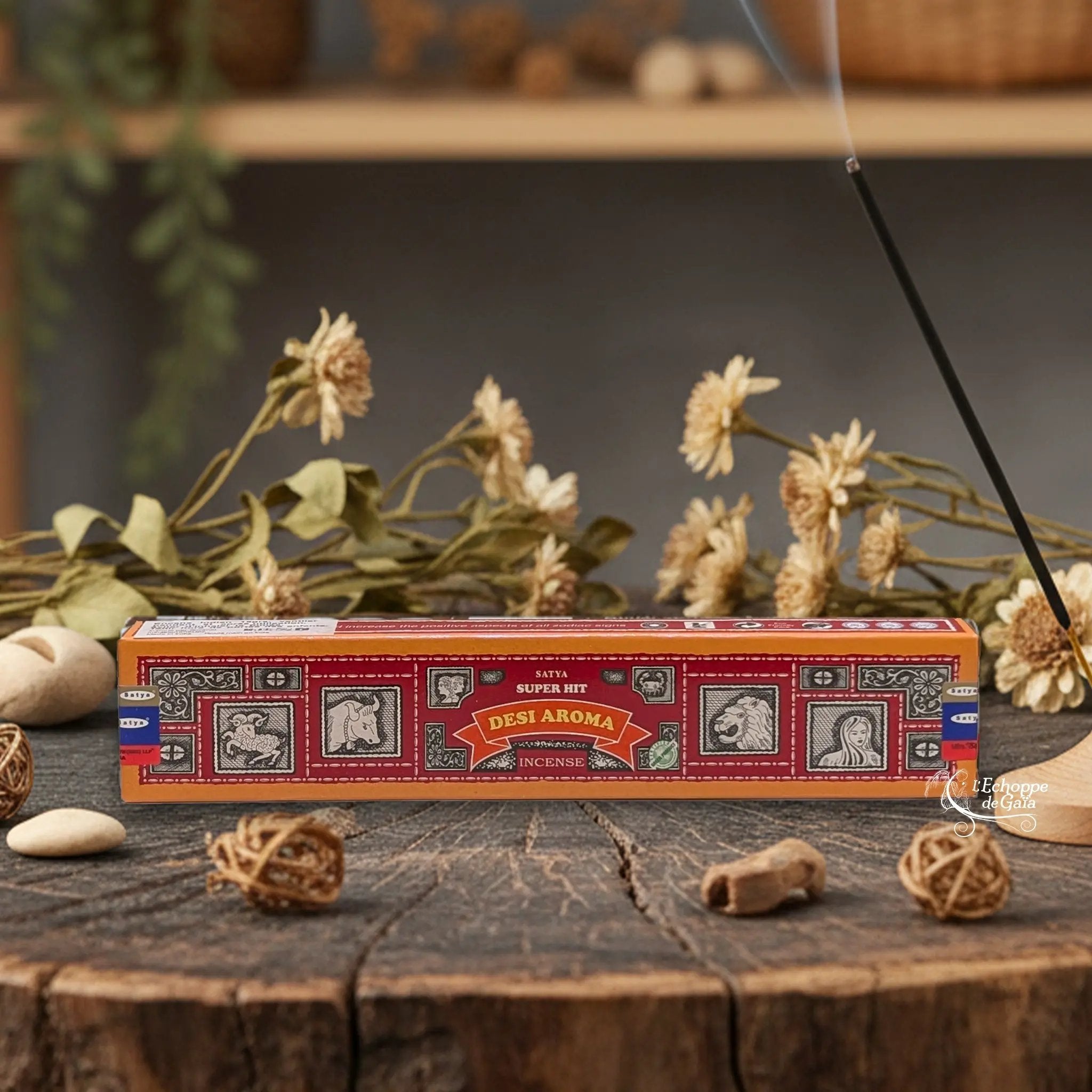Boîte d'encens Satya Super Hit "Desi Aroma" posée sur une table en bois avec des fleurs séchées et un bâton d'encens fumant.