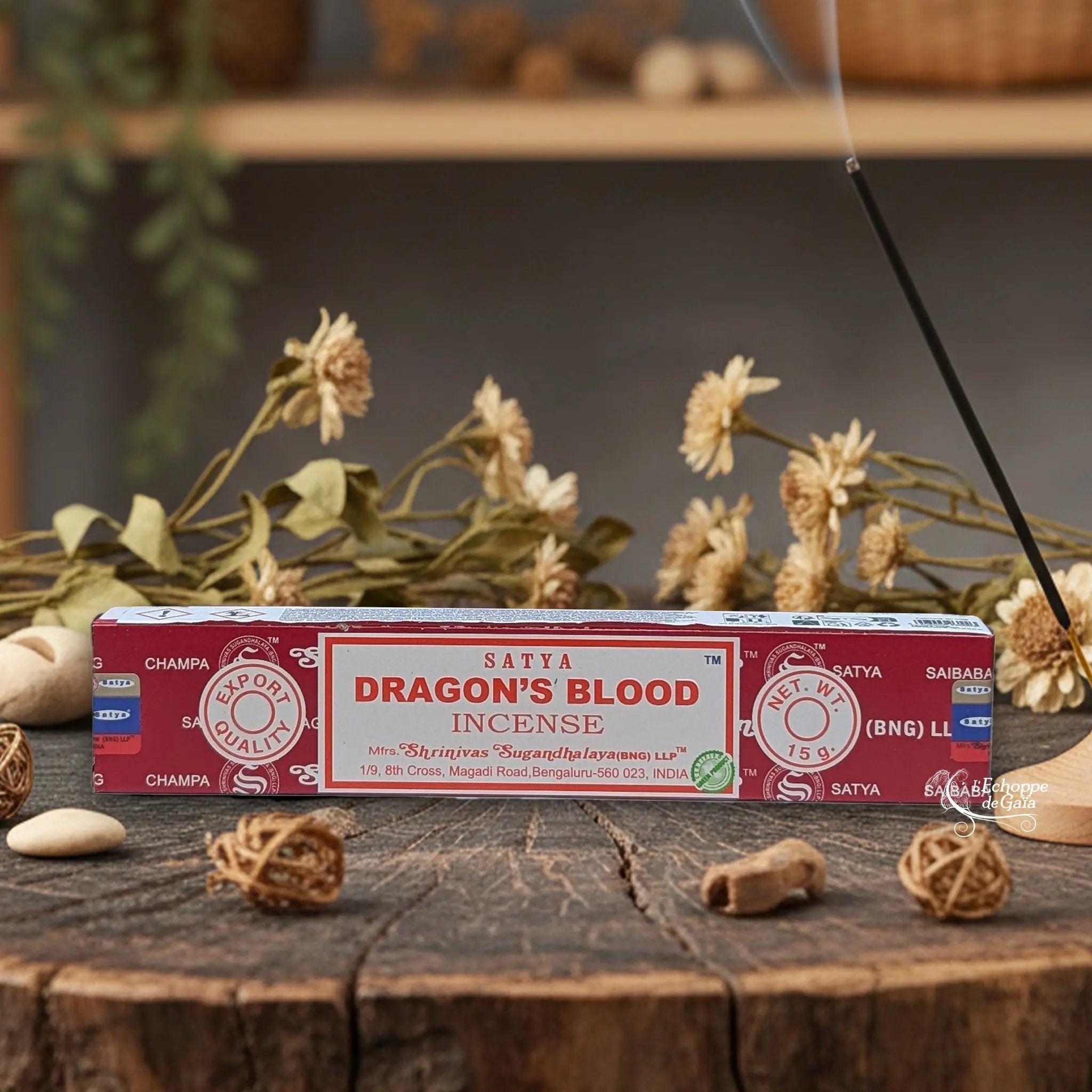 Boîte d'encens Satya "Dragon's Blood" posée sur une table en bois avec des fleurs séchées et un bâton d'encens fumant.
