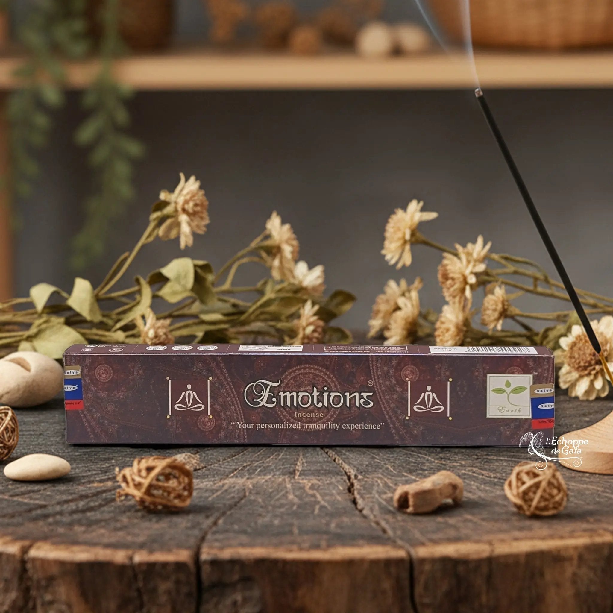 Boîte d'encens Satya "Emotions" posée sur une table en bois avec des fleurs séchées et un bâton d'encens fumant.